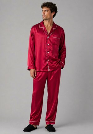 Homme portant un pyjama en satin rouge avec des passepoils blancs et des chaussons noirs, debout devant un fond gris uni, regardant sur le côté.