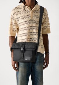 Borsa a tracolla nera con patta e chiusura a fibbia, dotata di una tracolla in tessuto strutturato e di una patch con logo. Indossata su una camicia beige a fantasia e jeans.