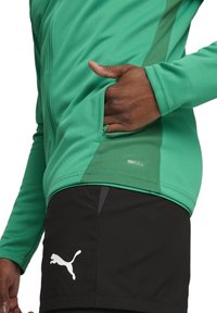 Groene sportjack met een ritszak en textuurpanelen aan de zijkanten, gecombineerd met zwarte shorts met een wit logo. Gemaakt van prestatie stof.