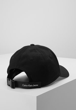 Czarna czapka baseballowa z regulowanym paskiem, ozdobiona białym napisem "Calvin Klein Jeans", prezentowana na białej powierzchni.