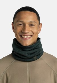 Dark green neck gaiter made of soft fabric, featuring a gathered design and a small branded label. Worn by a person in a beige top.