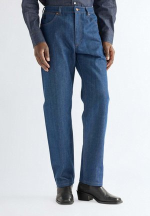 Mann trägt blaue Jeans mit geradem Bein, dunkles Hemd und schwarze Stiefeletten mit Absatz, steht vor einem einfarbigen Hintergrund.
