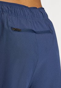 Κοντινή λήψη ναυτικού μπλε αθλητικών σορτς με μικρές περφορέτες και τσέπη με φερμουάρ στο πίσω μέρος της μέσης ενός ατόμου.