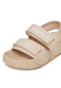 Beżowe sandały Reebok z dwoma regulowanymi paskami na rzep, teksturowaną podeszwą i wyściełaną wkładką, zaprojektowane dla komfortu i wygody noszenia.