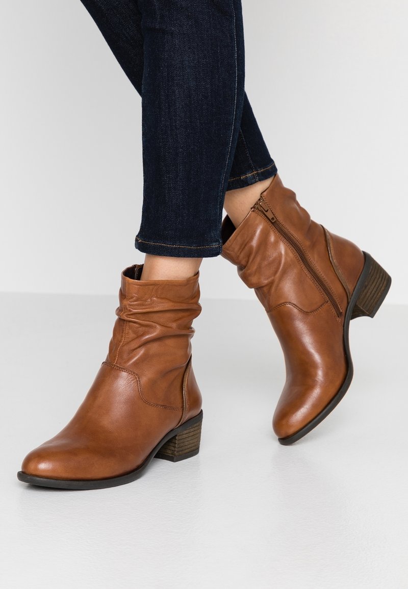 Braune Leder-Stiefeletten mit seitlichem Reißverschluss, getragen zu dunkelblauen Skinny-Jeans vor einem weißen Bodenhintergrund.