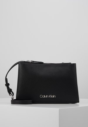 Zwart gestructureerd leren Calvin Klein polstasje met zilveren ritssluiting en logo, geplaatst op een wit oppervlak tegen een grijze achtergrond.