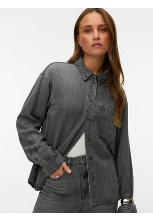 Frau trägt ein locker sitzendes dunkelgraues Jeanshemd über einem weißen Top, mit passenden grauen Jeans, Hand in der Tasche und langen braunen Haaren.