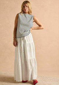 Prúžkovaná bezrukávová blúzka v námorníckej a bielej farbe, spárovaná s dlhou bielou bavlnenou sukňou s výšivkou a poschodovou konštrukciou, obutá s červenými topánkami.