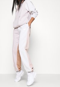 Nike-hoodie i mjuk rosa med en framficka och dragsko, parat med matchande byxor med vita och beige paneler; vita sneakers.