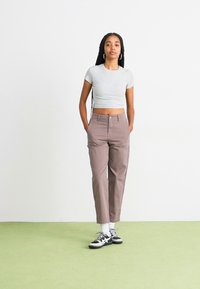 Grijze cropped t-shirt, beige rechte broek en zwarte sneakers met witte accenten. Achtergrond met een lichte muur en een groene vloer.