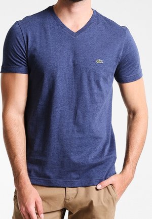 Mann trägt ein dunkelblaues V-Ausschnitt-T-Shirt mit kleinem grünem Krokodil-Logo auf der Brust und beigefarbenen Hosen, Hand in der Tasche.