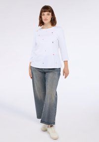 Blusa blanca de manga larga con pequeños acentos decorativos multicolores, combinada con jeans de mezclilla azul de pierna ancha y zapatillas de color claro.
