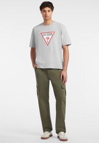 Camiseta de algodón gris con estampado de logo triangular rojo, combinada con pantalones cargo verde oliva, que cuentan con bolsillos laterales, y zapatillas blancas con detalles en marrón.