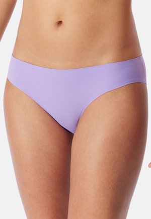 Frau trägt nahtlose fliederfarbene Bikini-Unterwäsche mit niedrigem Bund, zeigt Taille, Hüften und Oberschenkel vor einem einfarbigen Hintergrund.