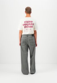Persona con cabello corto que lleva una camiseta blanca con el texto rojo "STUDIO ORIGINALS EDITION" y unos pantalones grises holgados, de pie contra un fondo blanco.