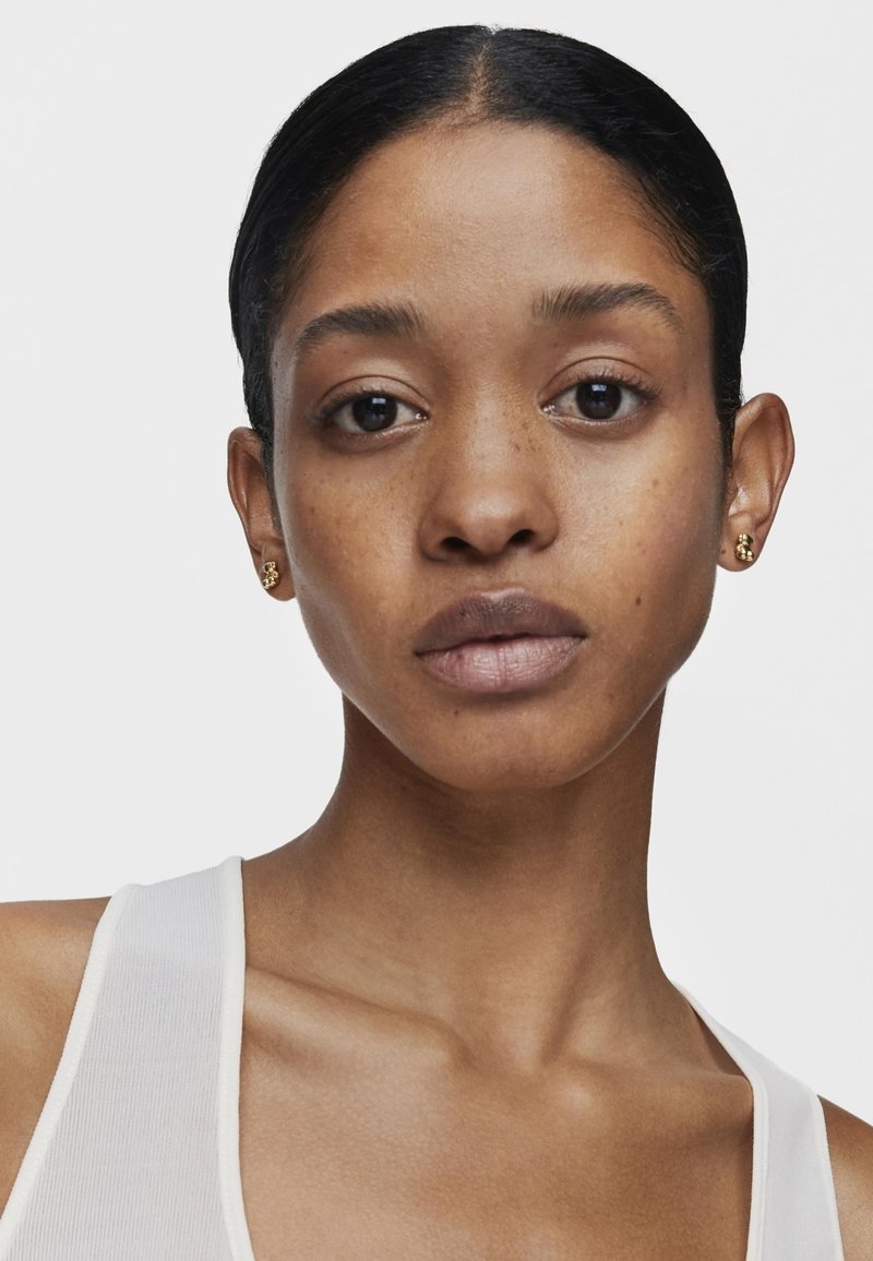 Pendientes de oro con una superficie texturizada. El modelo tiene un cutis suave y lleva una blusa sin mangas blanca, mostrando un aspecto natural.