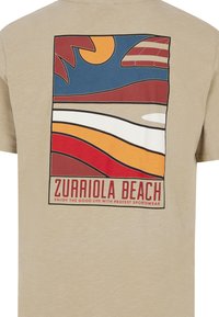 Camiseta de algodón beige con un diseño gráfico que presenta olas, sol y montañas en rojo, azul, naranja y crema. El texto dice "PLAYA ZURRIOLA."