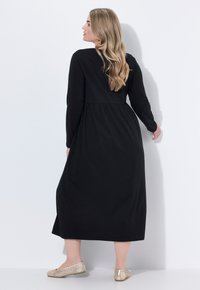 Robe noire à manches longues avec une taille froncée et une jupe fluide. Portée avec des ballerines tissées de couleur claire, vue de dos.