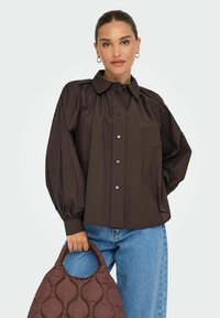 Camicia button-up marrone scuro con maniche a sbuffo, abbinata a una borsa marrone trapuntata. Donna che indossa jeans blu e orecchini a cerchio.