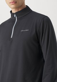 Fekete, hosszú ujjú sportos felső, félig cipzáros gallérral, világosszürke díszítéssel a cipzáron, és 'Calvin Klein' logóval a mellkason.