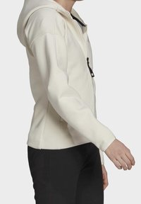 Světle krémová mikina na zip s přiléhavým střihem, žebrovanými akcenty na ramenou a hladkým povrchem. Obsahuje zip a černou táhlo.