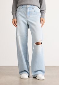 Pantalones de mezclilla azul claro con corte de pierna ancha, que presentan una rodilla desgastada, un diseño clásico de cinco bolsillos y costuras blancas.