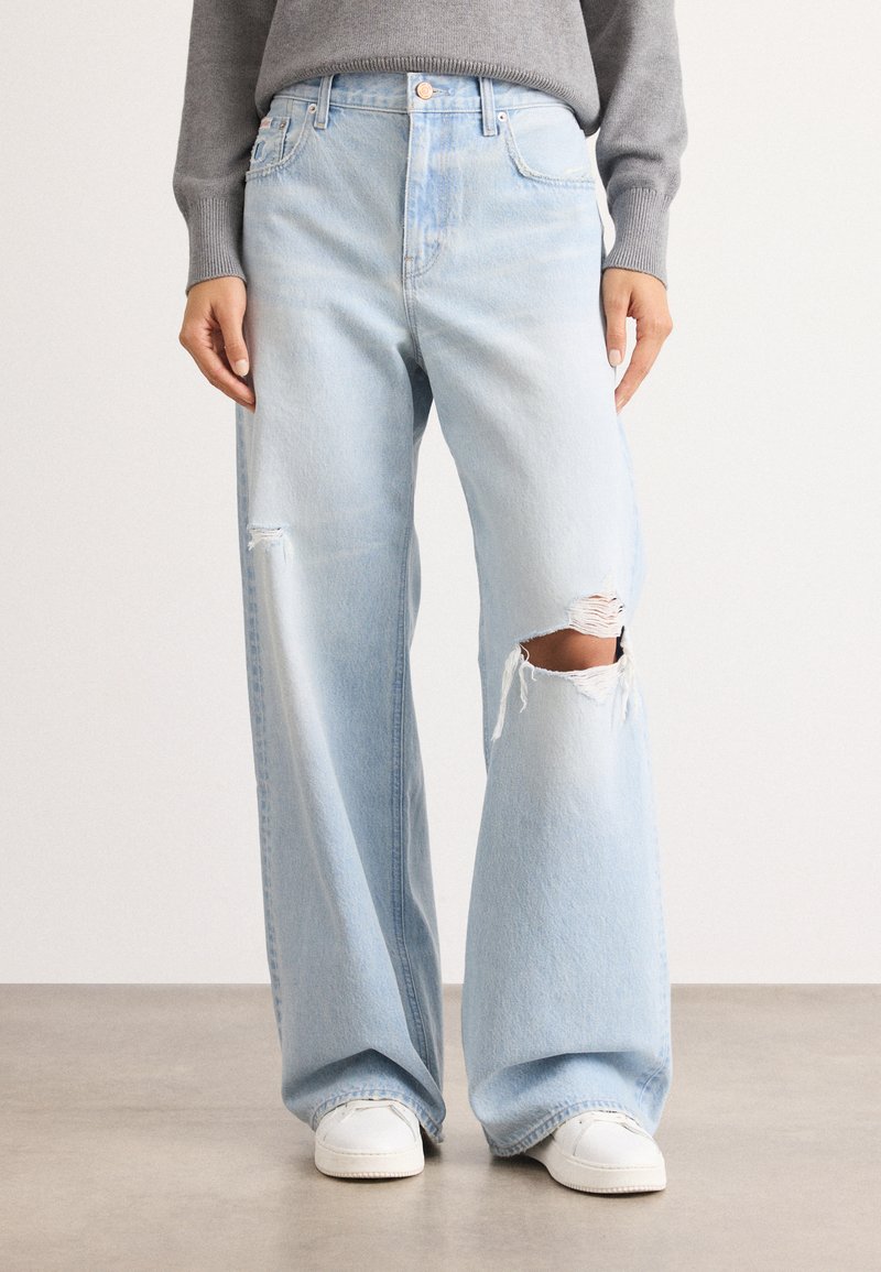 Pantalones de mezclilla azul claro con corte de pierna ancha, que presentan una rodilla desgastada, un diseño clásico de cinco bolsillos y costuras blancas.