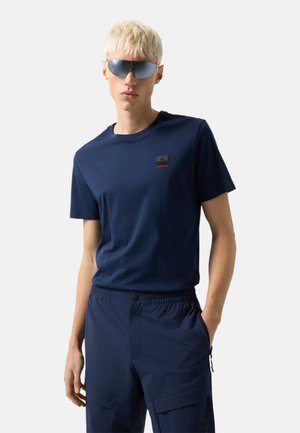 Blonde Person mit reflektierender Sonnenbrille, marineblauem Kurzarmhemd mit Logo-Patch und navyfarbenen Cargo-Hosen, die die Hände in den Taschen hat.