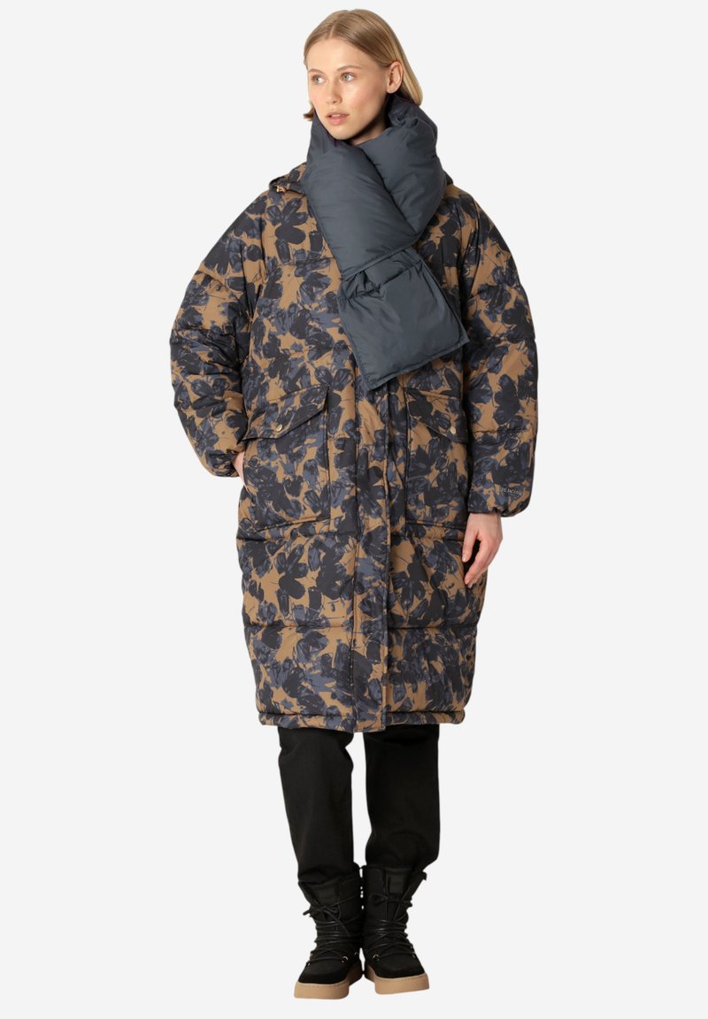 Abrigo acolchado con estampado floral oscuro en azul marino y marrón. Presenta una bufanda oscura desmontable, bolsillos frontales y una silueta relajada. Pantalones y botas negras.
