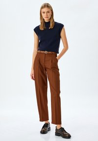 Marineblaues ärmelloses Top, kombiniert mit hochgeschnittenen, gefalteten braunen Hosen, ergänzt durch einen braunen Gürtel und dunkle Schnürschuhe.