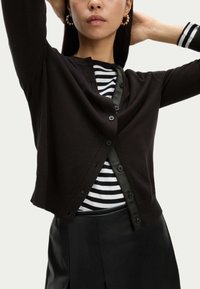 Cardigan noir avec un devant boutonné et des poignets côtelés sur un haut à rayures noir et blanc. Texture lisse, design simple et coupe ajustée.