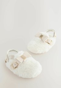 Balti pūkaini slippers ar platu dizainu, kuriem ir liels gaiši beidžu loks un zelta toņa sprādze augšējā daļā. Visā virsmā ir mīksta tekstūra.