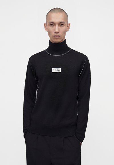 Homme portant un pull à col roulé noir avec une étiquette rectangulaire blanche sur la poitrine, debout devant un fond clair uni.
