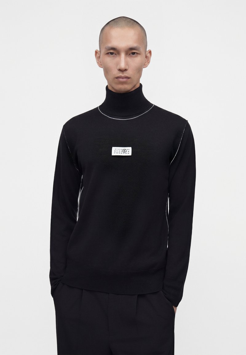 Homme portant un pull à col roulé noir avec une étiquette rectangulaire blanche sur la poitrine, debout devant un fond clair uni.