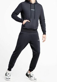 Felpa grigia scura con cappuccio, tasca frontale e polsini elasticizzati, abbinata a pantaloni jogger coordinati con caviglie a coste. Accenti del logo bianchi.