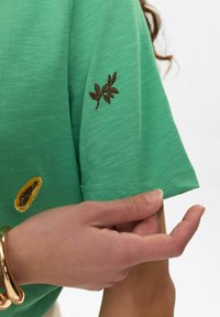 Mėtų žalia marškinėliai su trumpomis rankovėmis, tekstūruota medžiaga ir rudi išsiuvinėti lapų detalės ant rankovės bei geltonos vaisių motyvai šalia.