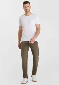 Homme avec les cheveux courts portant un T-shirt blanc avec une poche sur le devant, un pantalon vert olive et des baskets blanches, se tenant devant un fond clair.