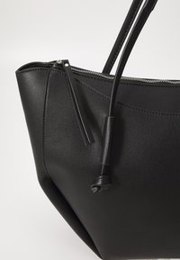 Sac cabas en cuir noir avec fermeture éclair, fines bretelles et texture lisse agrémentée de détails de surpiqûres subtils.