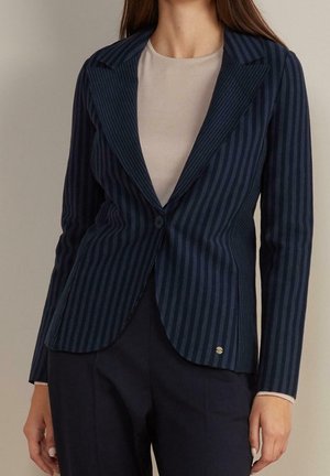 Femme portant un blazer bleu marine ajusté à fines rayures avec un seul bouton, par-dessus un top beige et un pantalon foncé.
