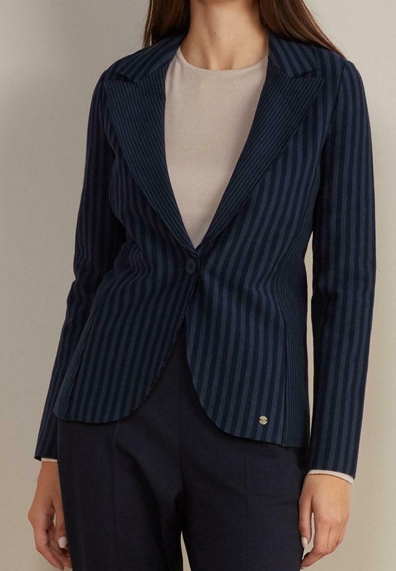 Femme portant un blazer bleu marine ajusté à fines rayures avec un seul bouton, par-dessus un top beige et un pantalon foncé.