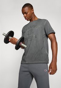 Camiseta de algodón gris con estampado de "Champion Training", combinada con pantalones de chándal grises. El sujeto sostiene un peso negro con mangos de metal.