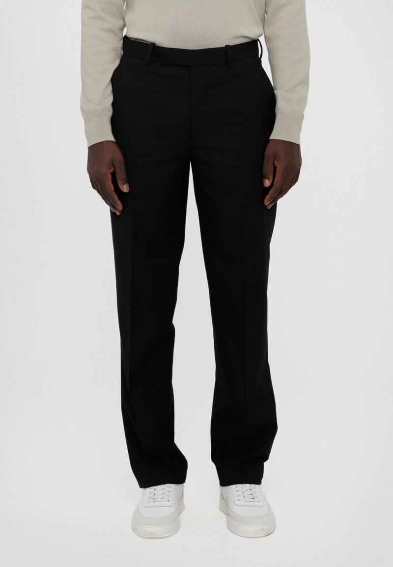 Schwarze Maßanzug-Hose aus glattem Stoff, mit geradem Bein und Gürtelschlaufen, kombiniert mit weißen Sneakern.