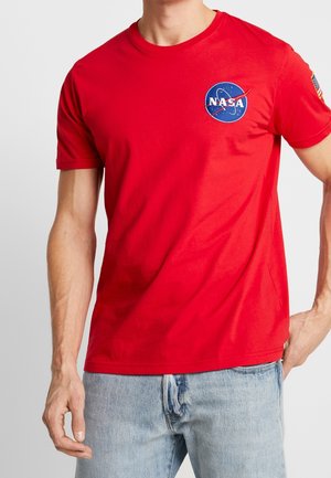 Mann in einem roten NASA-Logo-T-Shirt und hellblauen Jeans, steht mit einer Hand in der Tasche vor einem schlichten Hintergrund.