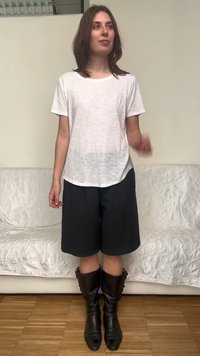 Jeune femme debout à l'intérieur portant un t-shirt blanc, un short noir jusqu'aux genoux et des bottes noires mi-mollet avec un canapé en arrière-plan.