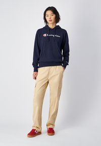 Felpa navy con logo bianco "Champion", tasca frontale, pantaloni cargo beige e sneaker in camoscio rosso con dettagli bianchi. Stile casual e vestibilità rilassata.