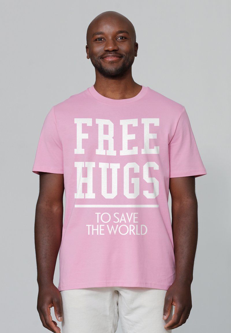 Lichtroze katoenen t-shirt met grote witte tekst "VRIJE KNUFFELS" en kleinere tekst "OM DE WERELD TE REDDEN." Korte mouwen, ronde hals.