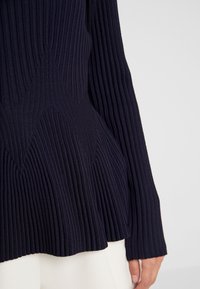 Tummansininen ribbineulottu neule, jossa on levenevä vyötärödetaili, jota käyttää vaaleat housut pukeva henkilö, kuvassa näkyy alavartalo ja käsivarsi.