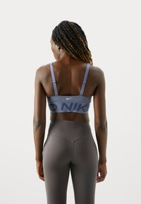Mujer con cabello trenzado que lleva puesto un sujetador deportivo azul de Nike y unas mallas de cintura alta gris oscuro, vista desde atrás contra un fondo plano.