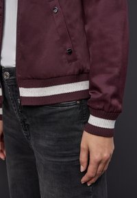 Veste bomber bordeaux avec une finition brillante, des poignets et un ourlet côtelés ornés de rayures blanches, assortie à un jean gris foncé.
