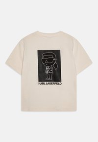 Bežs kokvilnas T-krekls ar melnu grafiku, kurā attēlota kartona figūra saules brillēs, zem kuras ir teksts "KARL LAGERFELD".